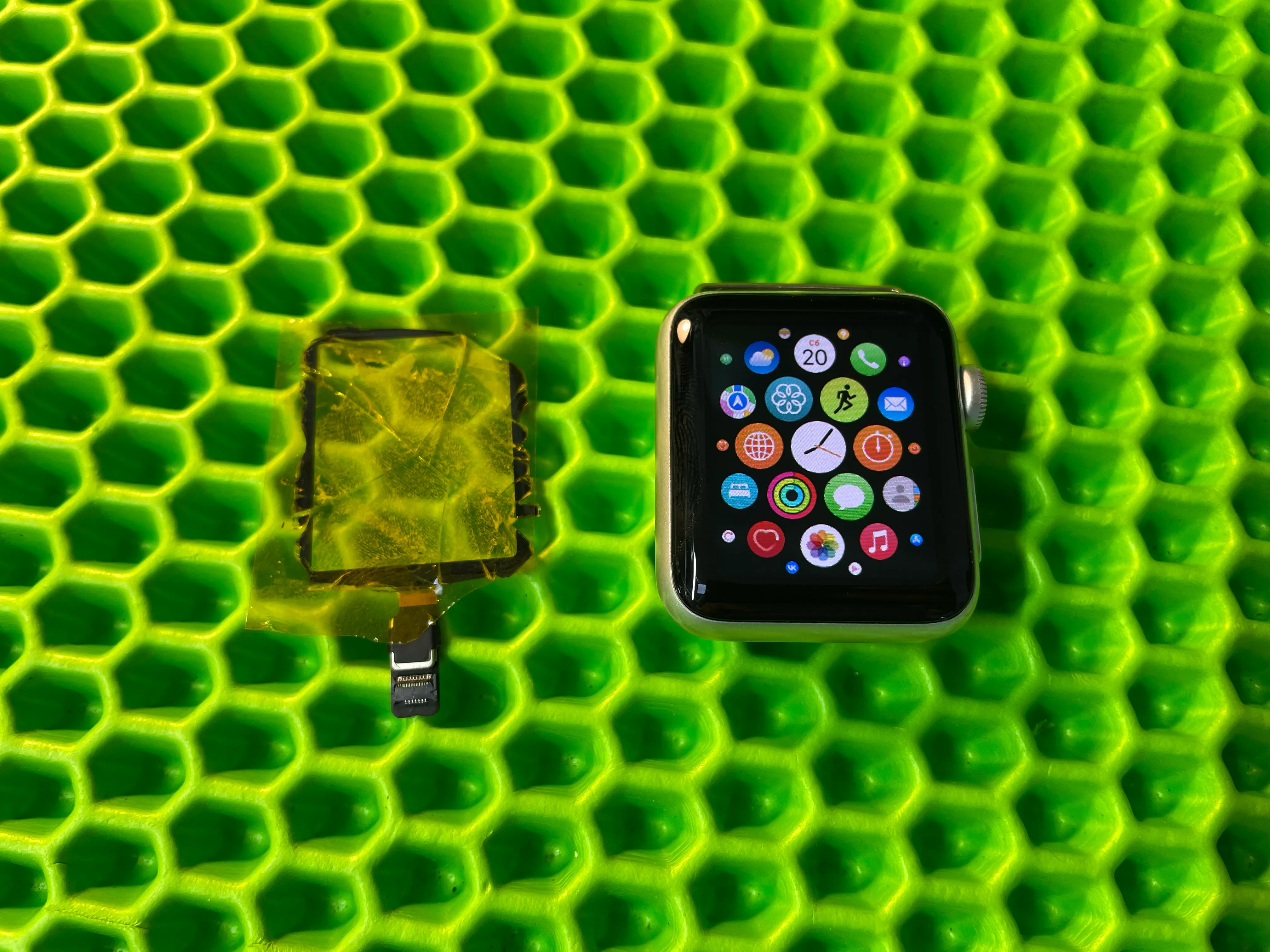 Apple Watch с экраном приложений рядом с повреждённым стеклом на зелёном коврике